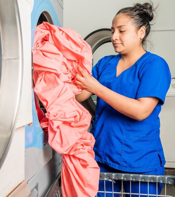 Vertical photo of a woman standing in front of a washing machine picking up a sheet in a laundry